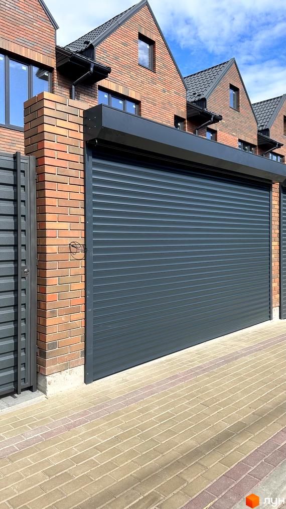 Житловий комплекс з темно-сірими ролетами на фасаді та цегляною кладкою. Бруківка на прибудинковій території.