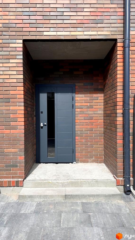 Вхідні двері сірого кольору з скляною вставкою в будинку з цегляною кладкою. Перед будинком бетонні сходи та плитка.