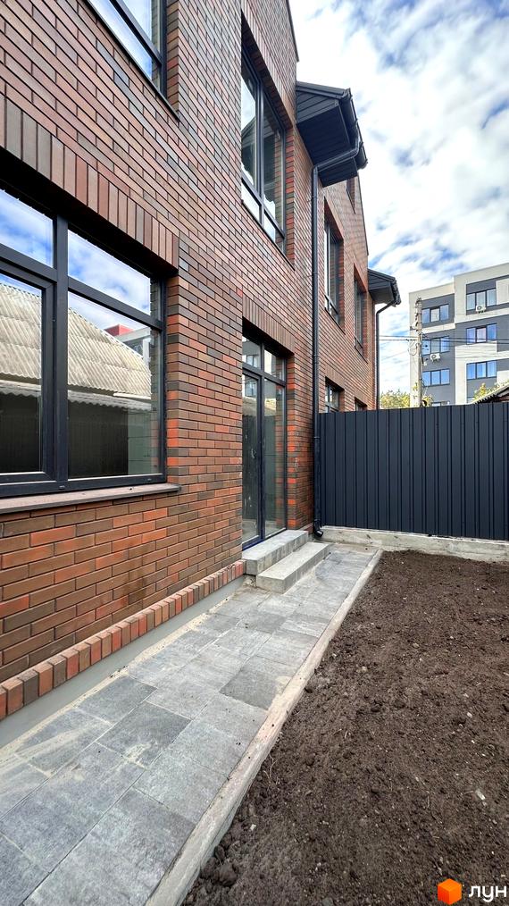 Цегляний будинок з темними вікнами та водостоками. Поруч вимощена доріжка та ділянка ґрунту.