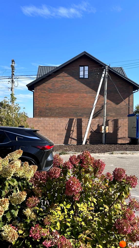 Цегляний будинок з темним дахом, біля якого ростуть квіти. Поруч з будинком стоїть чорний автомобіль та стовп з лініями електропередач.