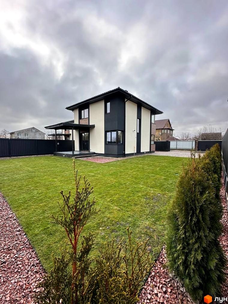 Будинок з біло-чорним фасадом та навісом біля входу. Зелений газон та туї на прибудинковій території.