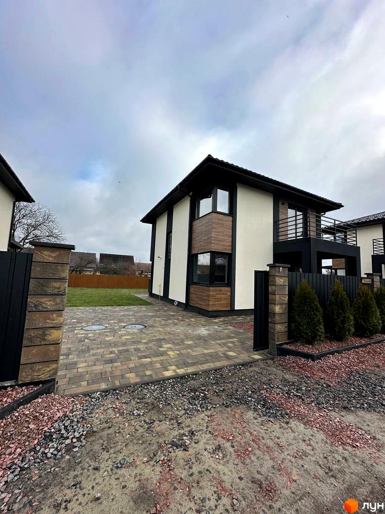 Сучасний двоповерховий будинок з дерев'яними елементами фасаду та балконом, розташований на вимощеній бруківкою ділянці з зеленим газоном та огорожею.