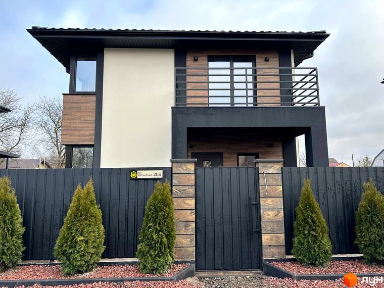 Фасад двоповерхового будинку з дерев'яними та білими вставками, балконом, чорним парканом та зеленими туями біля входу. Новобудова на вул. Яблунська, 20.