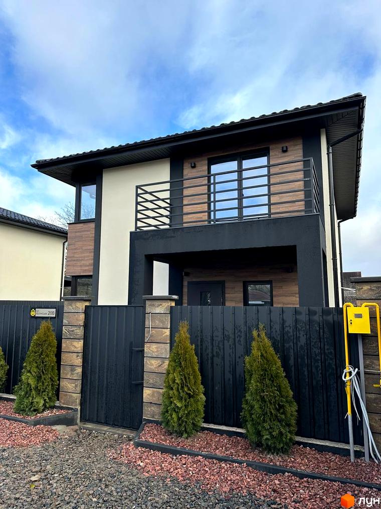 Сучасний двоповерховий будинок з дерев'яним оздобленням фасаду та балконом, огороджений парканом з туями та газоном.