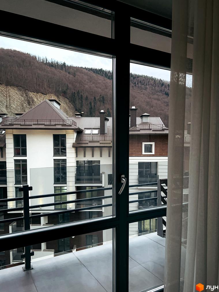 Вид з вікна на житловий комплекс Крайобраз з коричневими дахами та білими фасадами. Навколо будівель видно схил гори з деревами, що створює мальовничий краєвид.