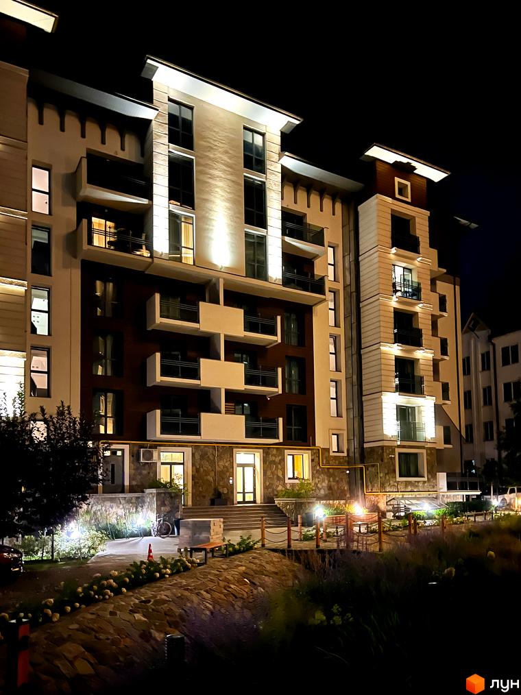 Житловий комплекс Крайобраз у вечірній час. Фасад будівлі оздоблений каменем та має багато балконів. На прибудинковій території розташовані ліхтарі та зелені насадження.