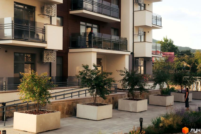 Зовнішній вигляд новобудови Крайобраз з балконами та кондиціонерами. Прибудинкова територія з деревами у великих бетонних горщиках та зеленню.