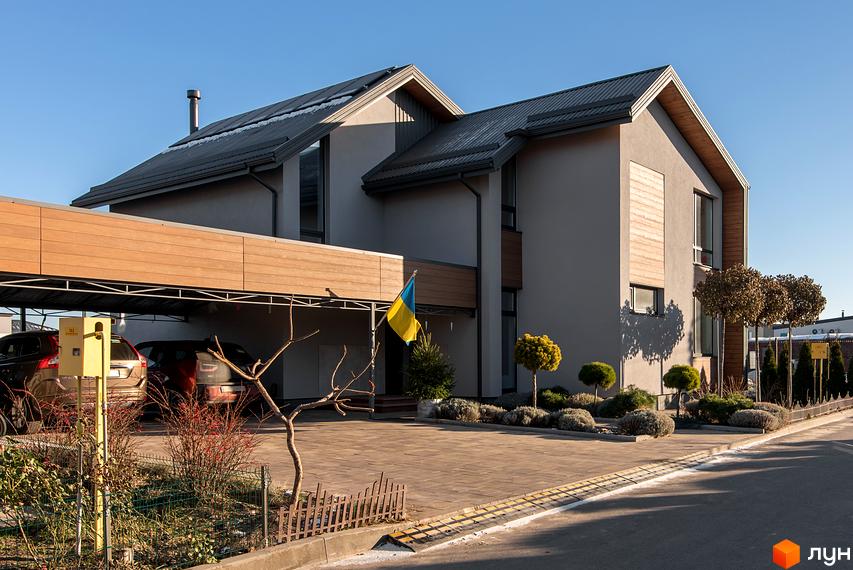 Котеджне містечко КМ Balaton Village. Зовнішній вигляд будинку з навісом для автомобілів та українським прапором. Прибудинкова територія з доглянутими кущами та деревами.