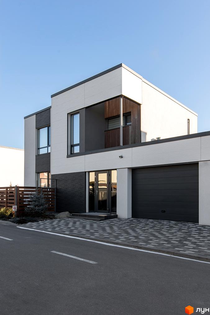 Котеджне містечко Balaton Village: будинок з біло-сірим фасадом, темними вікнами та гаражними воротами. Поруч вимощена ділянка та дерев'яний паркан.