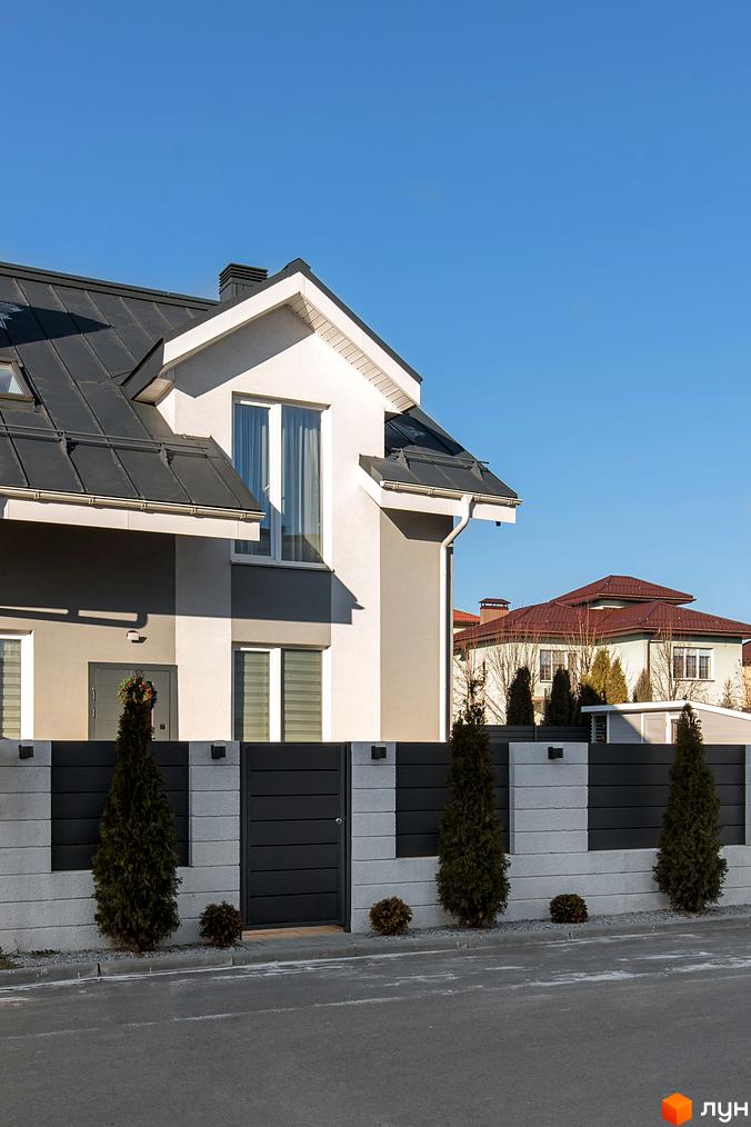 Котеджне містечко КМ Balaton Village: двоповерховий будинок з сірим дахом та біло-сірими стінами, огороджений парканом з туями. На задньому плані видно інший будинок з червоним дахом.