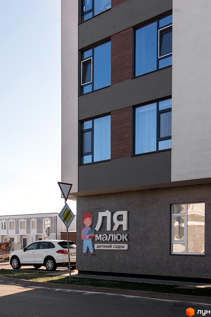 Зовнішній вигляд новобудови КМ Balaton Village з великими вікнами та фасадом з дерев'яними елементами. Поруч припаркований білий позашляховик та дорожні знаки.