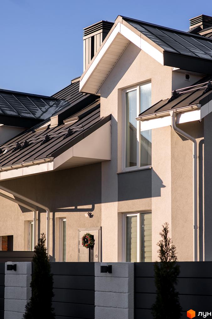 Котеджне містечко Balaton Village: фасад будинку з темним металевим дахом та білими вікнами. Поруч паркан з туями та ліхтарями.