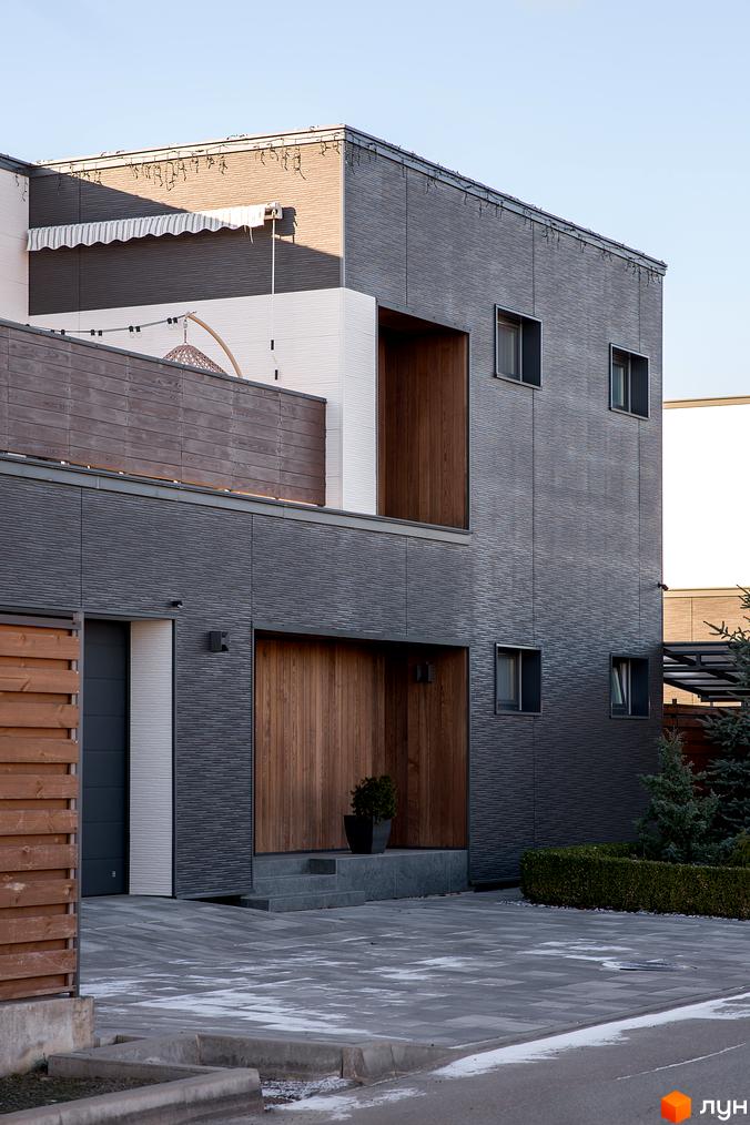 Котеджне містечко Balaton Village. Фасад будинку з темною текстурною плиткою та дерев'яними елементами, прибудинкова територія з бруківкою та зеленими насадженнями.