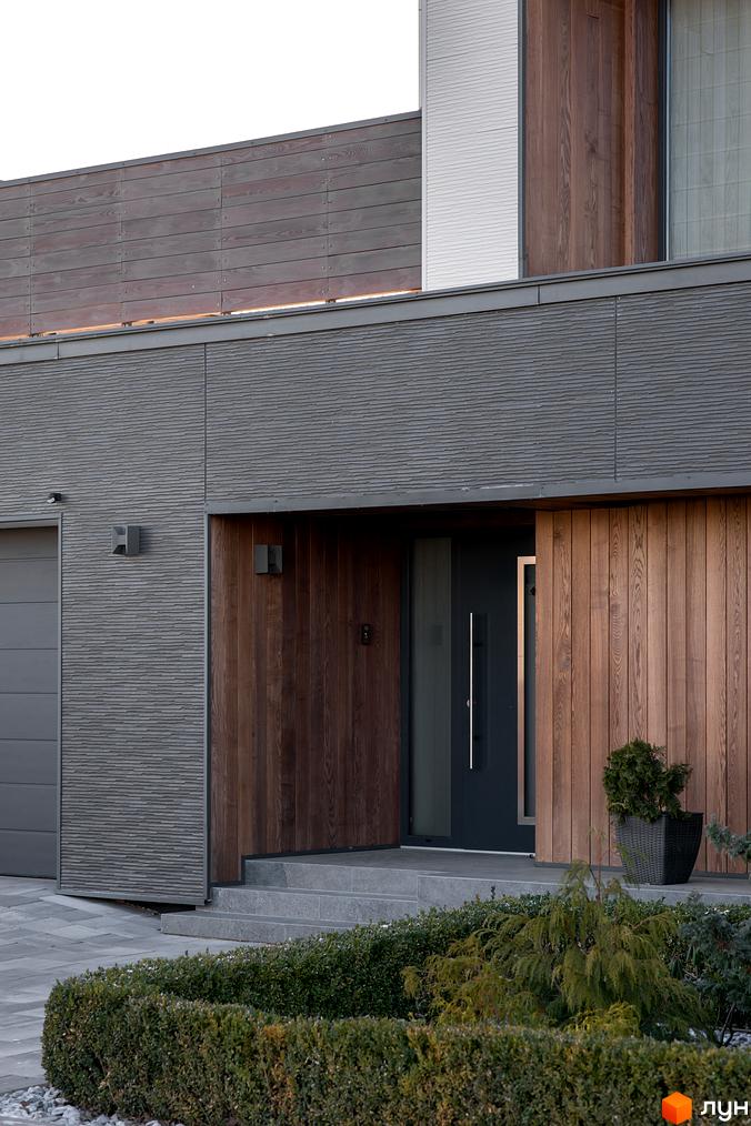 Котеджне містечко Balaton Village: фасад будинку з дерев'яними та сірими панелями, вхідна група з великими дверима та сходами. Навколо будинку зелені насадження та доріжки.