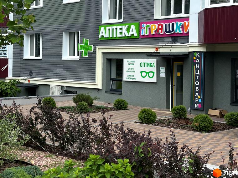 Фасад житлового комплексу Всі Свої з аптекою та магазином канцтоварів. Прибудинкова територія з кущами та бруківкою.