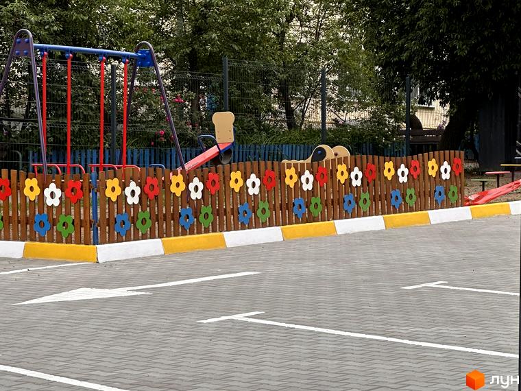 Дитячий майданчик з гойдалкою та гіркою, огороджений дерев'яним парканом з квітковими елементами. Територія біля новобудови ЖК Всі Свої.