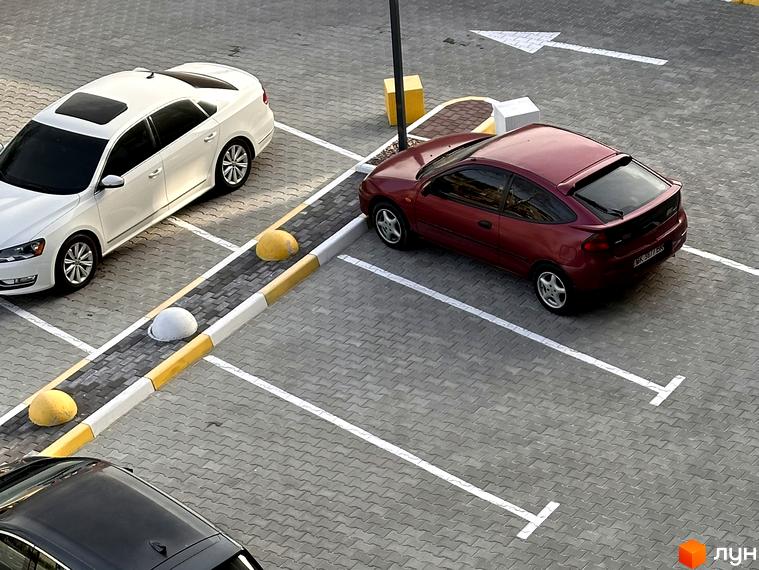 Паркінг біля житлового комплексу Всі Свої з білим седаном та червоним хетчбеком. Автомобілі припарковані на сірій бруківці.