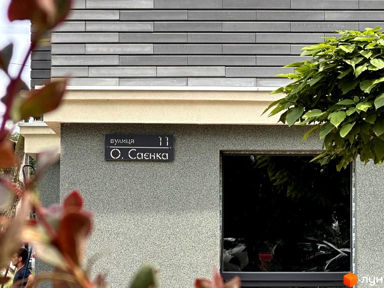 Зовнішній вигляд фасаду житлового комплексу Всі Свої з табличкою вулиці О. Саєнка, 11. Навколо будівлі зелені насадження та частина прибудинкової території.