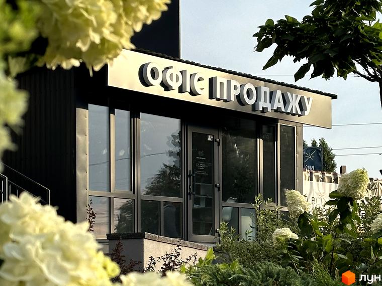 Зовнішній вигляд фасаду новобудови ЖК Всі Свої з великими вікнами та вивіскою. Навколо будівлі розташовані зелені насадження та квіти.