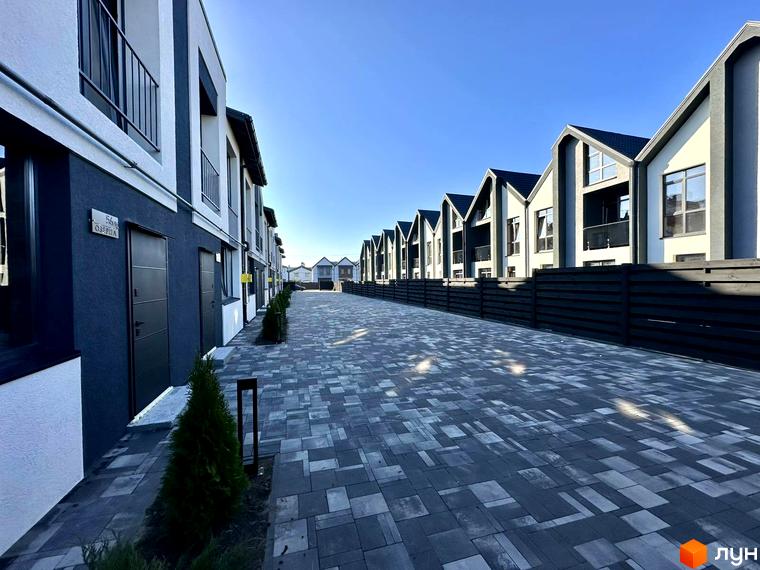 Таунхауси COMFORT RESIDENCE з сірими фасадами та чорними дахами, розташовані вздовж вимощеної сірою плиткою алеї з зеленими насадженнями.