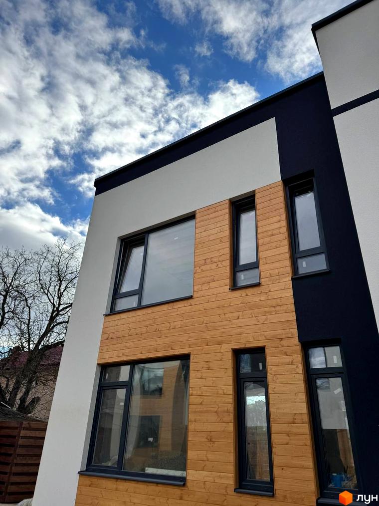 Котеджне містечко КМ Central Town: фасад будинку з дерев'яним оздобленням та великими вікнами на тлі хмарного неба. Поруч видно дерева та паркан.