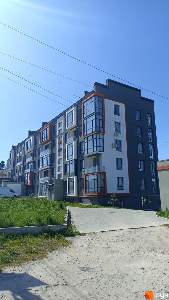 Зовнішній вигляд новобудови на вулиці Князя Володимира Великого, 8а, з сірими та білими фасадами та оранжевими акцентами. Навколо будинку розташована зелена зона та дорога.