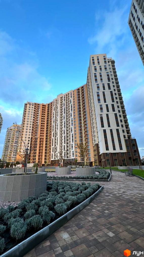 Новобудова KADORR City з вигнутим фасадом та балконами на тлі блакитного неба. Прибудинкова територія з доглянутими кущами та бруківкою.