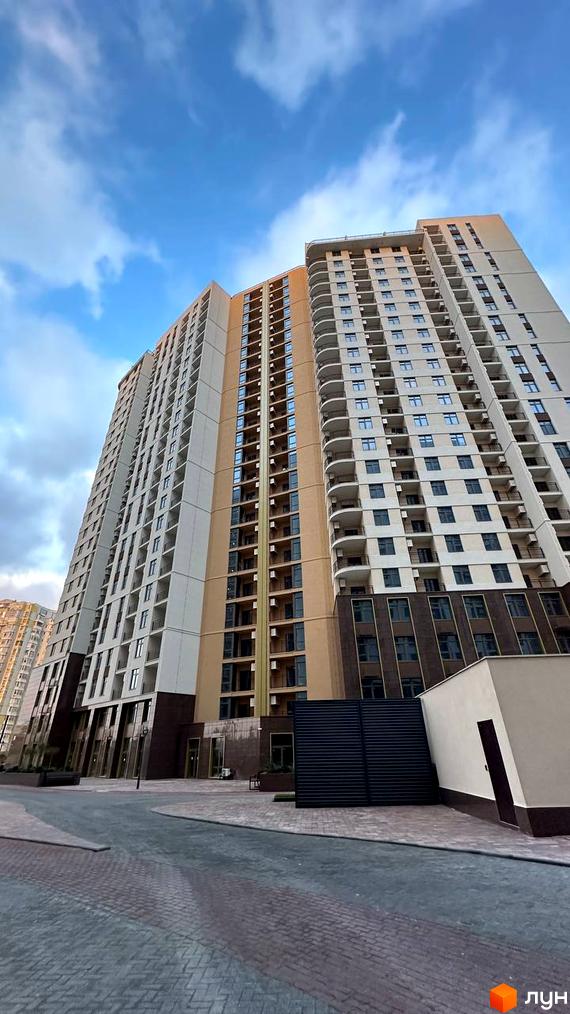 Високоповерховий житловий комплекс KADORR City з балконами та великою кількістю вікон на тлі блакитного неба. Прибудинкова територія з бруківкою та зеленими насадженнями.