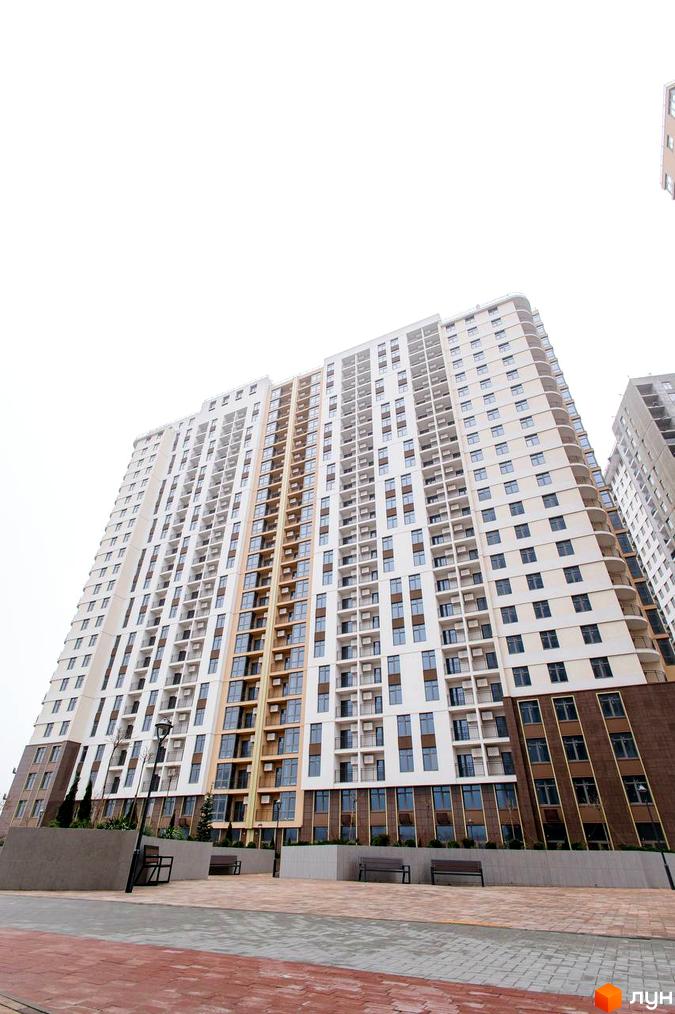 Високоповерховий житловий комплекс KADORR City з біло-бежевим фасадом та коричневими вставками. Поруч розташовані лавки та ліхтарні стовпи на бруківці.