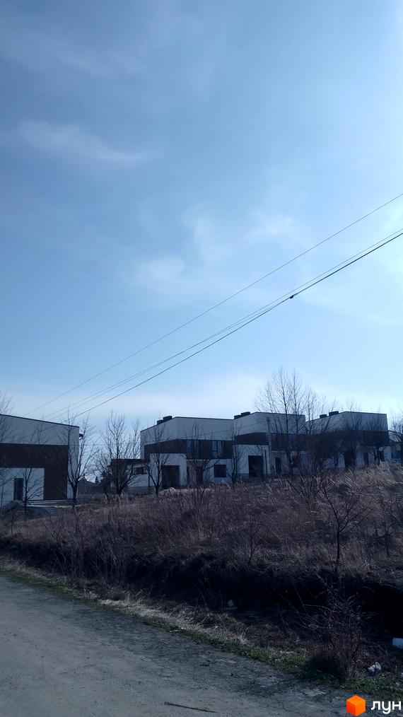 Зовнішній вигляд новобудови на вул. Вербицького з сірими та білими фасадами, оточеної деревами та сухою травою.