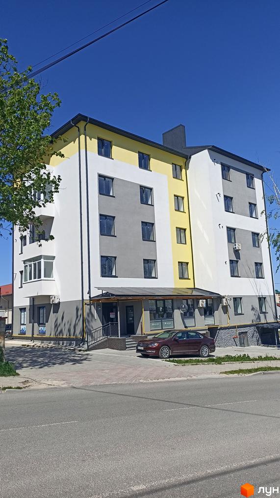 Новобудова на вулиці Незалежності з фасадом білого, сірого та жовтого кольорів. Перед будинком розташована прибудинкова територія з дорогою та припаркованим автомобілем.
