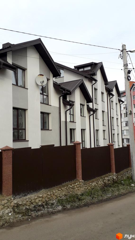 Зовнішній вигляд житлового комплексу на вулиці Квітки Цісик, 54а, б, в, г, з коричневим дахом та білими стінами, огородженого парканом. На прибудинковій території видно кам'яну огорожу та траву.