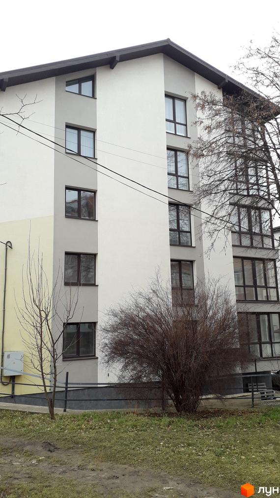 Зовнішній вигляд новобудови на вул. Квітки Цісик, 54а, б, в, г, з великою кількістю вікон та сірим фасадом. Прибудинкова територія з деревами та травою.