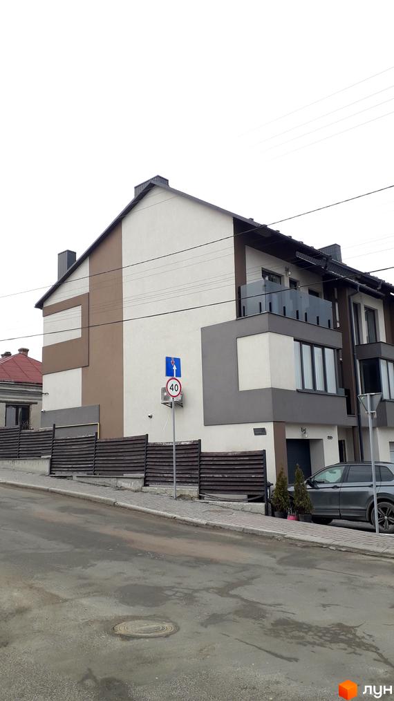 Фасад будинку з коричневими та сірими елементами, вікнами та балконами, на тлі дороги та паркану.