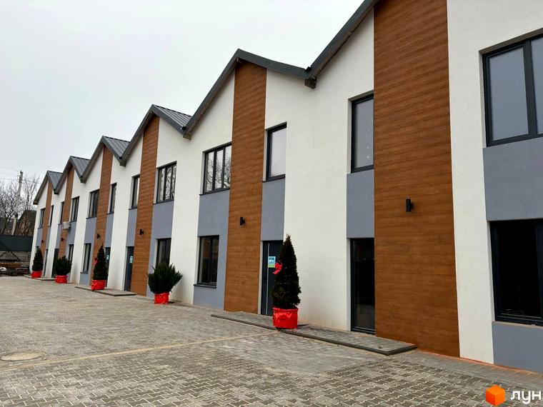 Таунхауси Klever House з білою та коричневою обшивкою, сірими вставками та темними вікнами, розташовані на бруківці з зеленими ялинками в червоних горщиках.