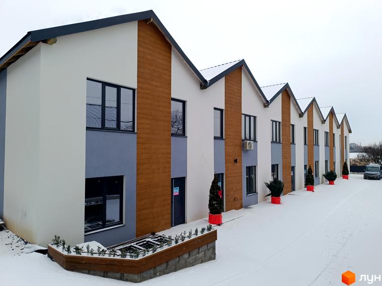 Таунхауси Klever House у біло-сірих кольорах з дерев'яними вставками під час снігопаду. Поруч розташовані червоні клумби з ялинками та парковка.