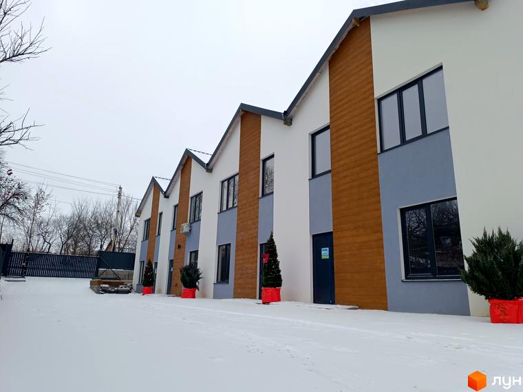 Таунхауси Klever House у зимовому ландшафті. Зовнішній вигляд будівель з дерев'яними та сірими елементами фасаду, вікнами та вхідними дверима. Прибудинкова територія засніжена, з ялинками у червоних горщиках.