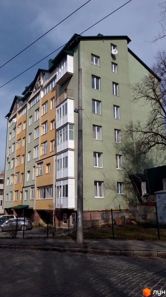 Фасад новобудови на вулиці Вояків Дивізії Галичина, 26, з різнокольоровими секціями та скляними балконами. Поруч розташована прибудинкова територія з парканом та деревами.