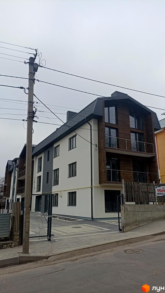 Зовнішній вигляд новобудови на вул. Весела, 46, з біло-сірими фасадами та дерев'яними вставками, огородженою прибудинковою територією та парканом.