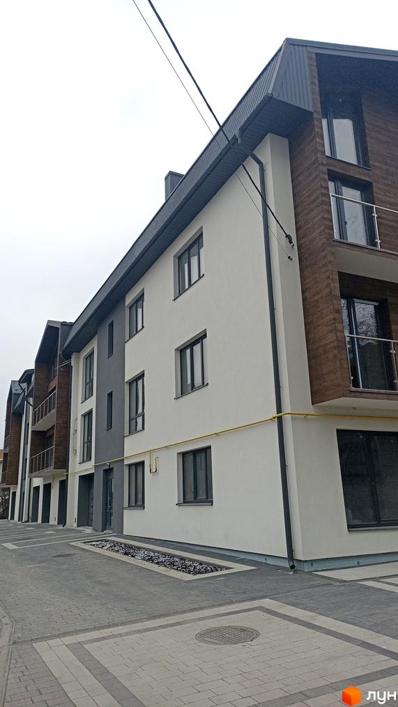 Фасад новобудови на вул. Весела, 46, з сірими та білими елементами та дерев'яними вставками. Прибудинкова територія вимощена плиткою.