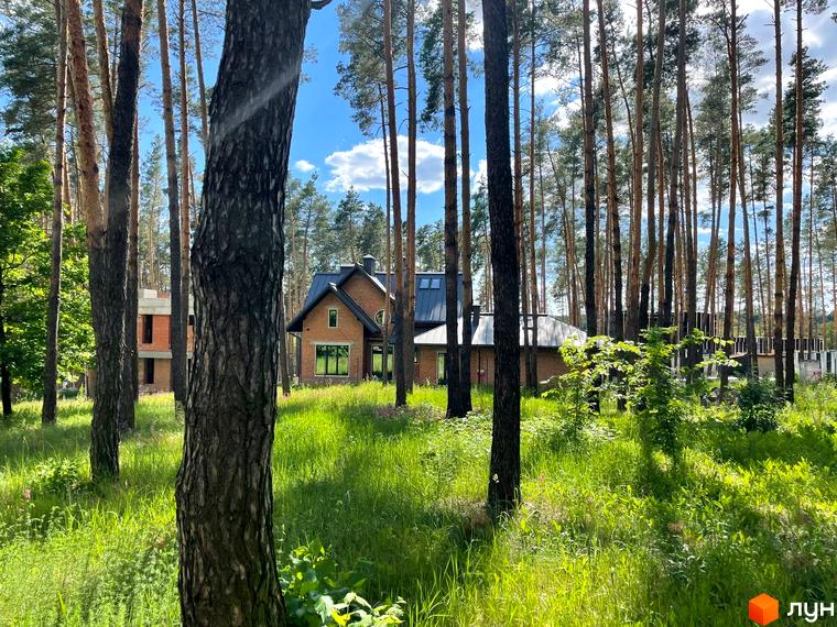 Котеджне містечко Swisstown. Цегляний будинок з темним дахом, розташований серед високих сосен. Навколо будинку зелена галявина з травою та кущами.