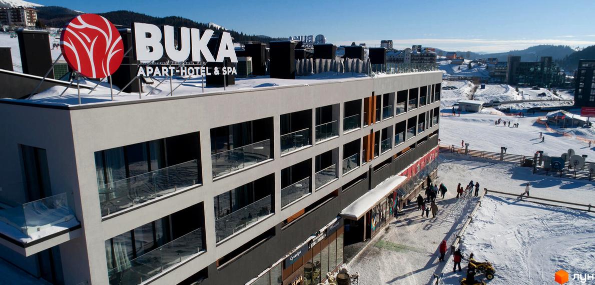 BUKA Apart-Hotel & SPA - це сучасний житловий комплекс з панорамними вікнами та балконами, розташований на фоні гірськолижного курорту з людьми, що катаються на лижах.