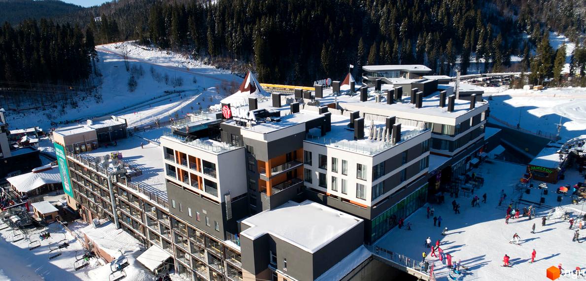 BUKA Apart-Hotel - це сучасний житловий комплекс з багатоповерховими будівлями та прилеглою територією для відпочинку та спорту. На зображенні видно будівлі комплексу, снігові схили та людей, що катаються на лижах.