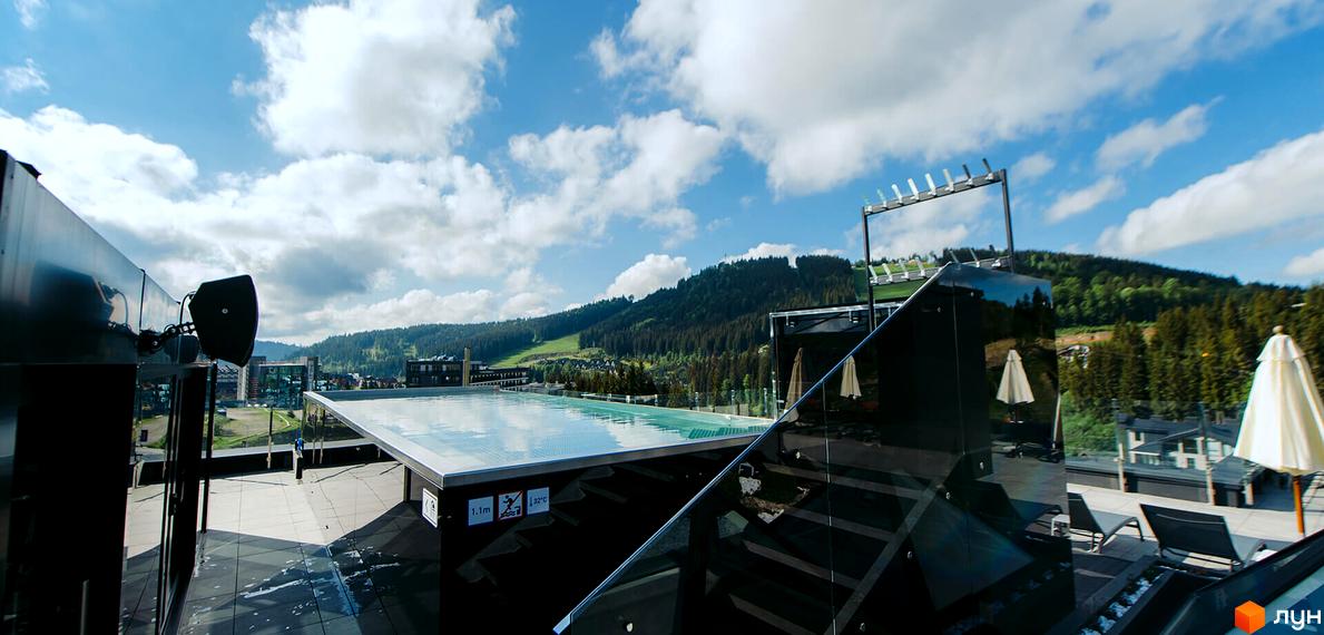 BUKA Apart-Hotel: панорамний вид на басейн на даху з видом на гори та ліси. Навколо будівлі розташовані шезлонги та парасольки.