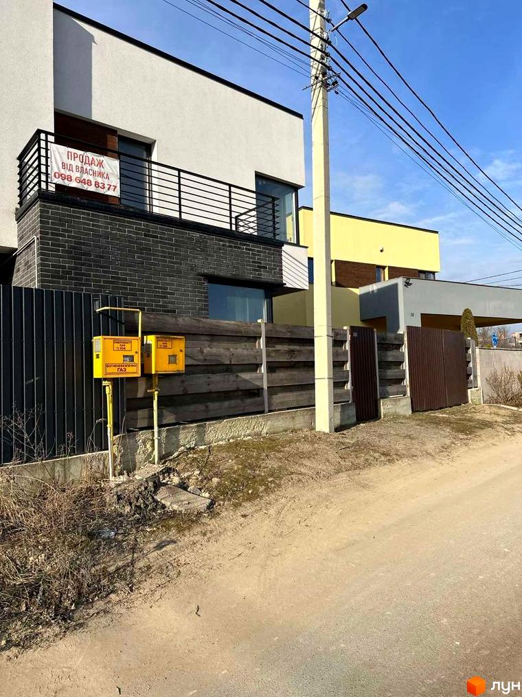 Будинок з білим та сірим фасадом, балконом та парканом. На прибудинковій території розташовані газові лічильники та дерева.