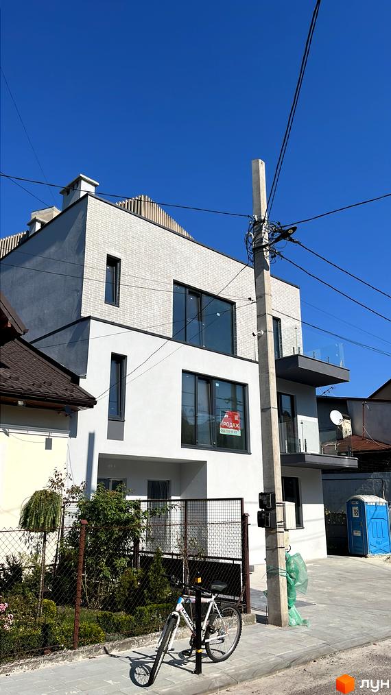 Зовнішній вигляд триповерхового будинку з балконом. Перед будинком велосипед та зелені насадження.