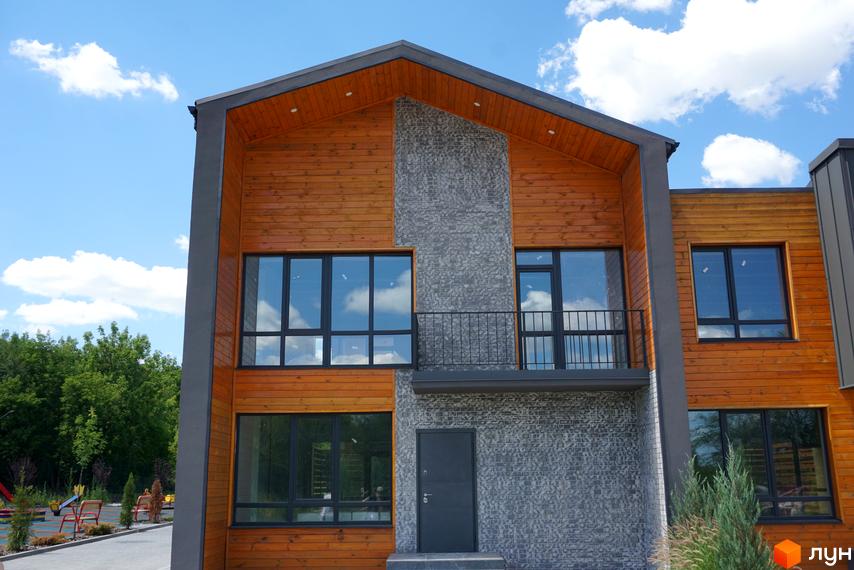 Котеджне містечко КМ ЛісоПаркове: фасад будинку з дерев'яною обшивкою та сірим оздобленням, великі вікна, балкон, двері. На задньому плані видно зелені дерева та дитячий майданчик.