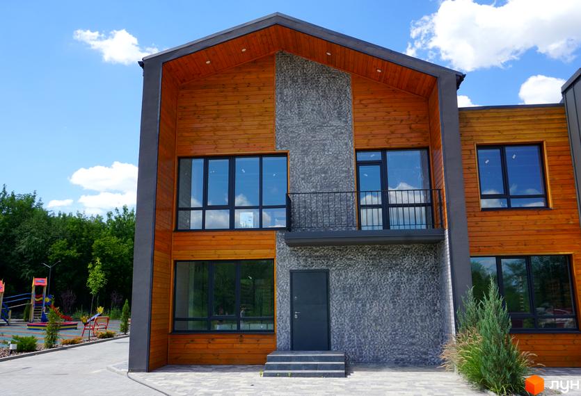 Котеджне містечко ЛісоПаркове. Зовнішній вигляд будинку з дерев'яним фасадом та сірою плиткою, вікнами та балконом. Поруч дитячий майданчик.
