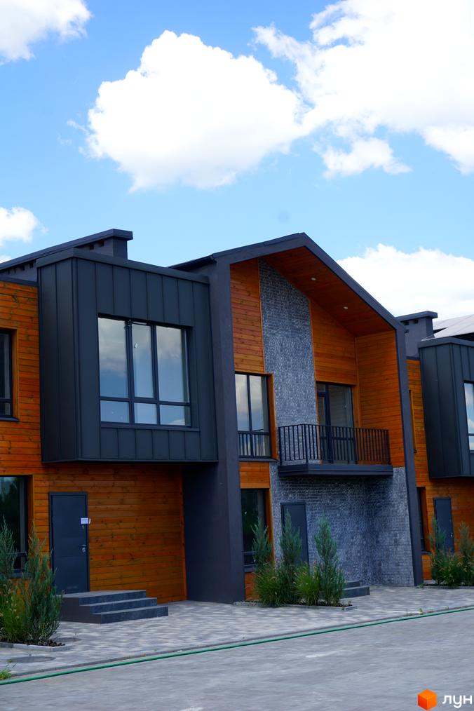 Котеджне містечко КМ ЛісоПаркове: фасади будинків з дерев'яними та сірими елементами, великі вікна, балкон, доглянута прибудинкова територія з зеленими насадженнями.