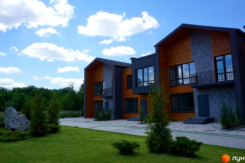 Котеджне містечко КМ ЛісоПаркове: фасади будинків з дерева та каменю, великі вікна, балкон, зелена галявина та дерева на території.